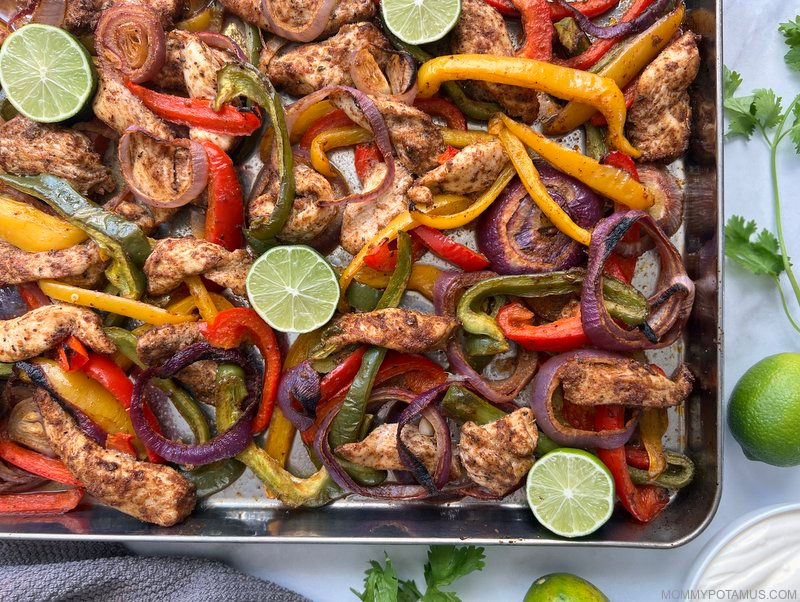 Chicken fajitas on sheet pan with lime wedges and sour cream on the counter nearby. 